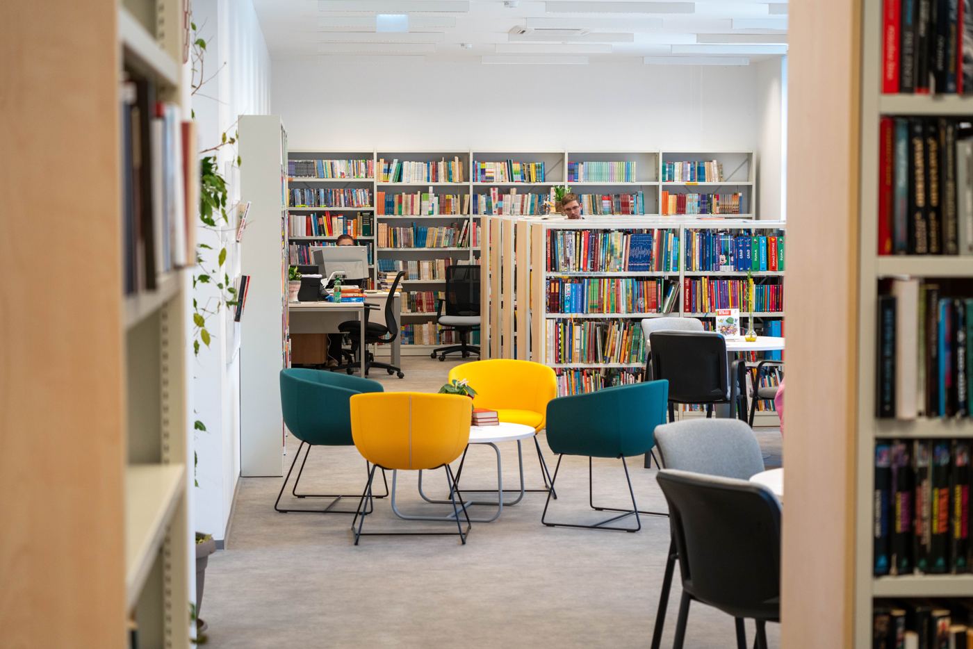 A bright public library reading area with teal and yellow chairs in front of full bookshelves.
